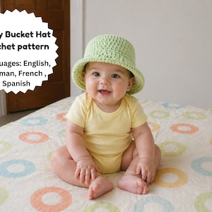 Könnte beinhalten: Ein Baby mit einem hellgrünen gehäkelten Fischerhut und einem gelben Strampler sitzt auf einer gemusterten Decke. Das Bild enthält den Text "Baby Bucket Hat crochet pattern" und listet Englisch, Deutsch, Französisch und Spanisch auf.