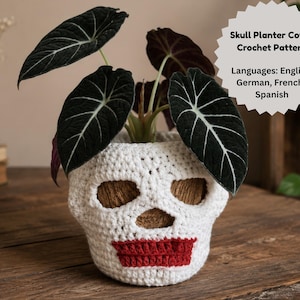 May include: A white crochet skull planter cover with red stitched lips, containing a dark green Alocasia plant. A circular tag displays the text: Skull Planter Cover Crochet Pattern. Languages: English, German, French, Spanish.