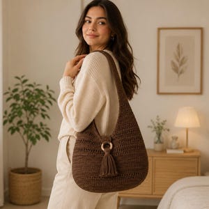 May include: A dark brown crochet hobo bag featuring a wide shoulder strap and a circular wooden ring accent at the centre front. A matching dark brown yarn tassel hangs from the wooden ring, adding texture to the solid, woven fabric construction.