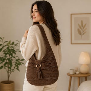May include: A dark brown crochet hobo bag featuring a wide shoulder strap, a circular wooden ring accent, and a matching yarn tassel. The bag has a textured, woven stitch pattern and a curved, slouchy silhouette that hangs from the shoulder.