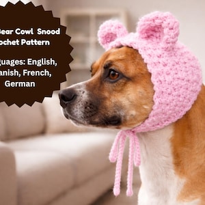 May include: A dog wearing a light pink crocheted bear cowl snood. The dog has brown and white fur. The snood has two small bear ears on top and ties under the chin. Text on the image reads "Dog Bear Cowl Snood Crochet Pattern".
