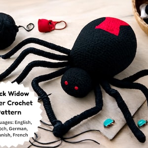 May include: A large, black crochet spider with a red hourglass marking on its back and red eyes. The spider has long, curved legs and is positioned on a wooden surface. The image also includes a ball of black yarn and the text "Black Widow Spider Crochet Pattern."