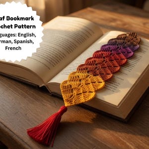 May include: A handmade crochet leaf bookmark in shades of yellow, orange, red, purple, and brown, with a red tassel, rests in an open book. The bookmark is made of yarn and has a detailed leaf pattern. The image includes text that says "Leaf Bookmark Crochet Pattern".