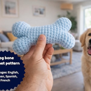 Puede incluir: Un juguete con forma de hueso para perros, de color azul claro y tejido a ganchillo, se sostiene en la mano de una persona. El juguete tiene forma de hueso y está hecho de hilo suave. Un perro golden retriever está en el fondo. La imagen incluye el texto "Dog bone crochet pattern".