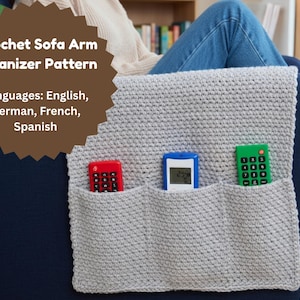 Op de afbeelding: Een lichtgrijze gehaakte sofa-arm organizer met drie vakken, met een rode, blauwe en groene afstandsbediening. De tekst op de afbeelding luidt "Crochet Sofa Arm Organizer Pattern" en vermeldt Engels, Duits, Frans en Spaans als beschikbare talen.