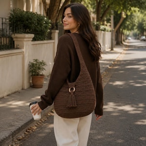 May include: A dark brown crochet hobo bag featuring a wide shoulder strap, a circular wooden ring connector, and a matching tassel detail. The bag has a textured, woven pattern and is worn over the shoulder, resting against the side of the body.