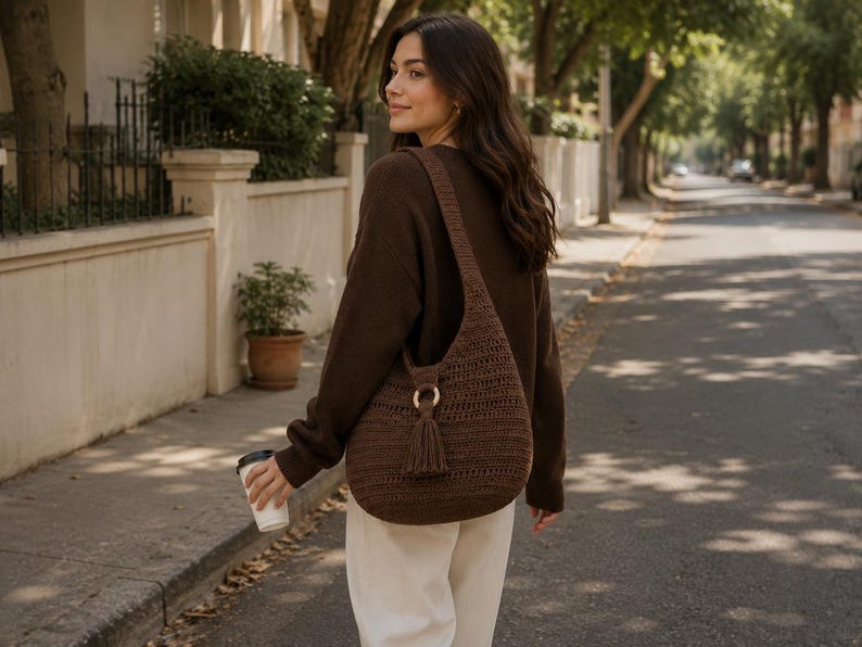 May include: A dark brown crochet hobo bag featuring a wide shoulder strap, a circular wooden ring connector, and a matching tassel detail. The bag has a textured, woven pattern and is worn over the shoulder, resting against the side of the body.