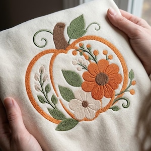 May include: A cream-colored sweatshirt featuring an embroidered design of a pumpkin outlined in orange thread. The pumpkin is decorated with stitched orange and cream flowers, green leaves, and brown stems, creating a detailed botanical motif.