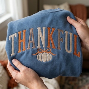 May include: A blue crewneck sweatshirt featuring the word THANKFUL embroidered in an arched pattern with alternating beige and orange letters. Below the text is an embroidered beige pumpkin with an orange bow tied around its stem.