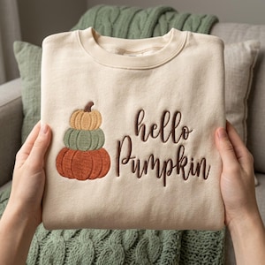 Può includere: Una felpa girocollo color crema con un ricamo raffigurante tre zucche impilate nei colori giallo, verde salvia e arancione bruciato. A destra delle zucche, il testo hello pumpkin è ricamato con un carattere corsivo marrone scuro.