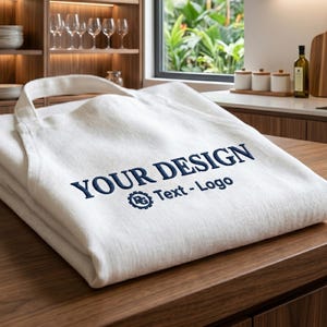 Könnte beinhalten: Eine gefaltete cremeweiße Küchenschürze mit dunkelblauer Stickerei, die YOUR DESIGN Text - Logo liest, sowie ein kleines kreisförmiges Zahnradsymbol mit den Buchstaben RG, das unter dem Haupttext auf der Vorderseite des Stoffes platziert ist.