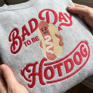 May include: A light gray sweatshirt featuring red embroidered text that reads "BAD DAY TO BE A HOTDOG". The design includes a central embroidered graphic of a sad-faced hotdog in a bun with a white squiggle of mustard, rendered in brown, tan, and white.