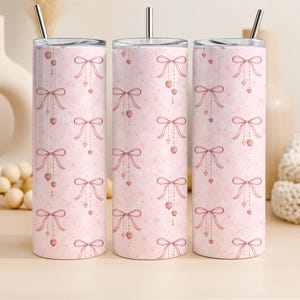 Puede incluir: Tres vasos térmicos rosas con un patrón repetido de lazos, corazones y fresas. Cada vaso tiene una pajita y tapa plateadas. El fondo es rosa claro con pequeños corazones y estrellas.