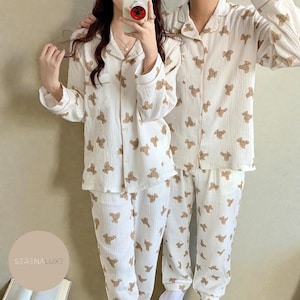 May include: Matching white pajama sets patterned with brown teddy bears. Each set includes a button-down shirt and trousers. The pajamas have a relaxed fit and are made of a soft fabric.