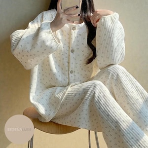 Könnte beinhalten: Cremefarbenes Pyjama-Set mit floralem Muster. Das Set beinhaltet ein Button-Down-Oberteil mit rundem Kragen und langen, puffärmeligen Ärmeln sowie eine passende Hose. Der Stoff wirkt gesteppt und weich, perfekt zum Entspannen.