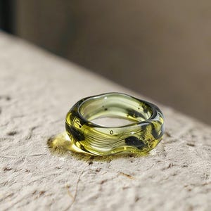 Könnte beinhalten: Ein transparenter, olivgrüner Ring mit einem wellenförmigen Design. Der Ring hat eine abgerundete Form und scheint aus Glas oder einem ähnlichen Material zu bestehen. Der Ring liegt auf einer strukturierten, hellfarbigen Oberfläche.