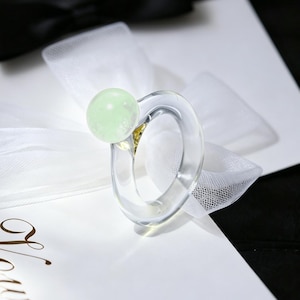 May include: A clear, circular ring featuring a single, smooth, pale mint-green spherical bead. The ring is positioned on a white card with the word Vows printed in elegant script, accented by a white tulle bow and a black satin bow.