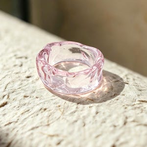 May include: A translucent light pink ring crafted from smooth glass. The band features an irregular, wavy, and organic shape with a glossy finish that catches the light, creating subtle reflections across its curved and rounded surface.
