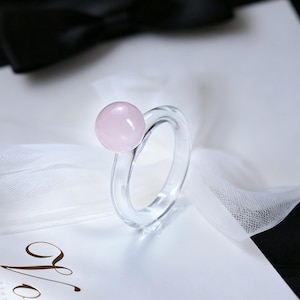 May include: A clear, transparent ring featuring a single, smooth, round, light pink bead attached to the top of the band. The ring is constructed from a solid, glass-like material, showcasing a simple, circular design with a polished finish.