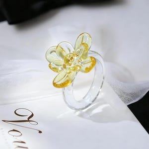 May include: A clear glass ring with a yellow flower design. The flower has multiple petals and a yellow center. The ring is sitting on a white surface.