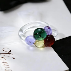 Puede incluir: Un anillo transparente con un grupo de cuentas de vidrio de colores. Las cuentas son en tonos de verde, morado, amarillo y rojo. El anillo está sobre una superficie blanca.