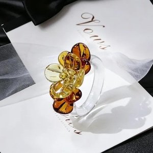 May include: A clear glass ring with a cluster of amber-colored flower petals. The petals vary in shade from light yellow to deep brown. The ring is set on a white card with the word "Vows" in elegant script.