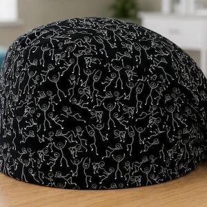 May include: Black surgical scrub cap featuring a repeating white line drawing pattern of stick figures. The cap is tied in a bow at the back. The cap is on a light brown wooden surface.