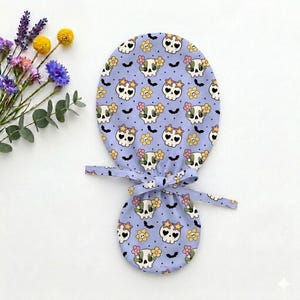 May include: A light purple fabric scrub cap featuring a repeating pattern of white skulls with heart-shaped eyes, yellow star-shaped glasses, and pink and yellow flowers, interspersed with small black bats and tiny dark purple dots.
