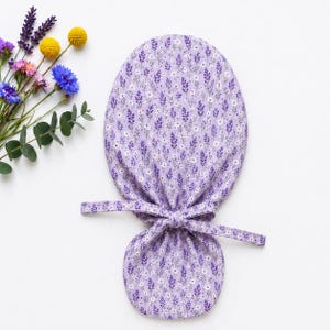 May include: A light purple fabric scrub cap featuring a repeating pattern of small lavender sprigs and tiny white flowers. The cap has a rounded top section and a gathered lower section secured with two fabric ties tied into a simple knot.