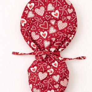 May include: Red fabric scrub cap with a pattern of white hearts, bows, and polka dots. The cap has a drawstring closure and is designed for medical professionals or those in the healthcare field. The fabric is a vibrant red.