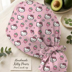 Könnte beinhalten: Rosa OP-Haube mit einem sich wiederholenden Muster aus Hello Kitty-Gesichtern, Erdbeeren und Blumen. Die Haube hat eine Schleife und den Text "Handmade Kitty Power Power scrub cap".