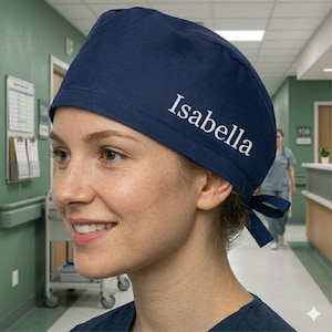以下が含まれることがあります： 白で「Isabella」と刺繍された、濃いネイビーブルーの手術用スクラブキャップ。キャップは、お揃いの濃いネイビーブルーのリボンで後ろで結ばれています。背景には病院の廊下が写っています。