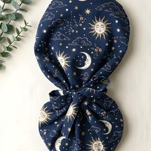 May include: A dark blue fabric scrub cap with a celestial design featuring suns, moons, stars, clouds, and floral accents. The cap is tied with a matching bow. The overall theme is celestial and whimsical.