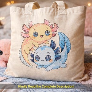May include: Beige tote bag featuring an embroidered design of two cartoon axolotls. One axolotl is yellow with pink gills, and the other is blue. The text "Kindly Read the Complete Description!" is at the bottom of the bag.