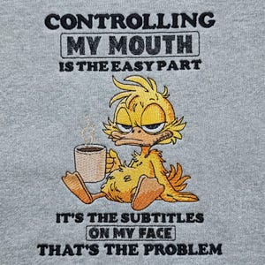 May include: A heather gray fabric featuring an embroidered yellow duck holding a steaming mug. The text reads: CONTROLLING MY MOUTH IS THE EASY PART. IT'S THE SUBTITLES ON MY FACE THAT'S THE PROBLEM. The duck has a grumpy expression and orange feet.