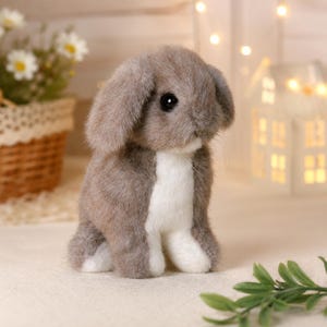 Può includere: Un coniglio di peluche con morbida pelliccia marrone chiaro e petto e zampe bianche. Il coniglio ha lunghe orecchie pendenti, occhi rotondi scuri e un piccolo naso. È raffigurato in posizione seduta, mostrando una consistenza realistica e dettagli.