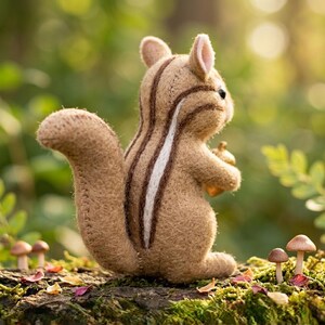 May include: A handcrafted plush chipmunk toy, in shades of brown with white and dark brown stripes, holding a small acorn. The chipmunk is positioned on a mossy log with miniature mushrooms, set against a blurred green background.
