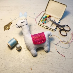 May include: A handmade white felt llama with a pink saddle, blue ears, and black stitched details. Sewing notions include a spool of blue thread, a thimble, scissors, pins, and a gold-colored charm. The items are arranged on a white wooden surface.