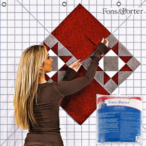 May include: A white Fons & Porter design wall featuring a printed grid pattern with diagonal lines. The wall is displayed with a red and grey patterned quilt block arrangement and includes a blue and white product packaging bag with text.