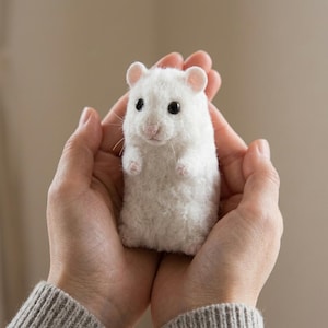 May include: A small, white, needle-felted hamster figurine with a fuzzy texture, round black eyes, and a light pink nose. The hamster is depicted in a standing position with tiny front paws visible, showcasing detailed craftsmanship.