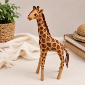 Peut inclure: Une figurine de girafe debout fabriquée en feutrine, présentant un corps beige avec des taches brun foncé, une crinière brun foncé le long du cou et une queue fine avec une extrémité touffue. La girafe a de petites oreilles et des yeux sombres.