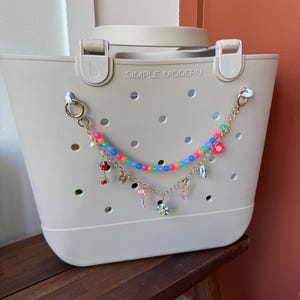 Può includere: Una borsa tote grigio chiaro con le parole "SIMPLE MODERN" stampate sul davanti. Una catena di perline con perline e ciondoli colorati è appesa sul davanti della borsa. I ciondoli includono un fungo, un fenicottero e un fiore.