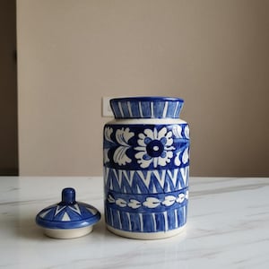 Puede incluir: Un tarro de cerámica azul y blanco con tapa, con un patrón floral y geométrico. El tarro es cilíndrico, con la tapa colocada al lado. El diseño está pintado a mano, mostrando una estética tradicional. El tarro está sobre una superficie blanca.