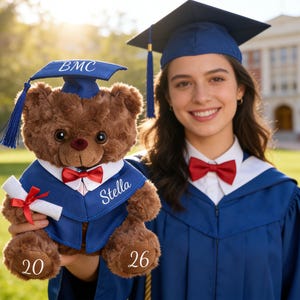 May include: A person in a blue graduation gown and bow tie holds a brown teddy bear wearing a graduation cap and gown. The bear has the name "Stella" on its gown and the numbers "20" and "26" on its paws.