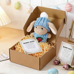 Puede incluir: Un conejito de peluche con gorro y suéter de punto azul con diseño de fresa, metido en una caja de cartón llena de paja. La caja incluye una tarjeta que dice "Bunny Hugs". Huevos de Pascua y flores decoran el fondo.