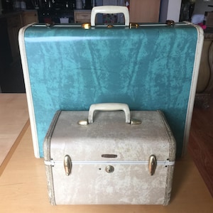 May include: A large teal suitcase with a marbled texture and a cream-colored handle, positioned behind a smaller, rectangular cream-colored vanity case. The vanity case features a central lock, two gold-toned latches, and a matching cream handle.