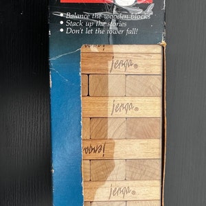 Könnte beinhalten: Eine blaue Schachtel mit einem weißen und roten Jenga-Spiellogo. Die Schachtel zeigt eine Nahaufnahme der Holzblöcke, die zu einem Turm gestapelt sind. Der Text auf der Schachtel lautet "A Milton Bradley Game" und "Jenga".