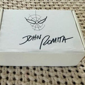 May include: A white cardboard box with a black Spider-Man logo and the signature "JOHN ROMITA." The box is rectangular and sits on a textured, beige surface. The Spider-Man logo features a stylized spider web design.