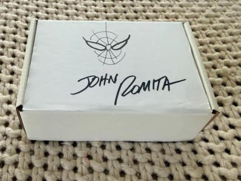 May include: A white cardboard box with a black Spider-Man logo and the signature "JOHN ROMITA." The box is rectangular and sits on a textured, beige surface. The Spider-Man logo features a stylized spider web design.