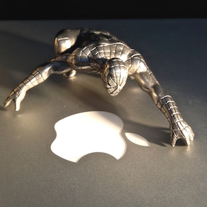 May include: A silver-colored Spider-Man figurine in a crouching pose. The figurine is detailed with textured lines and is positioned over a white Apple logo on a gray surface. The lighting creates shadows, highlighting the figure's form.